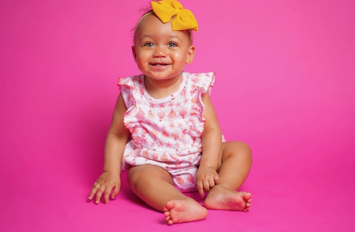 Smiling baby girl with a yellow bow, seated on a pink backdrop